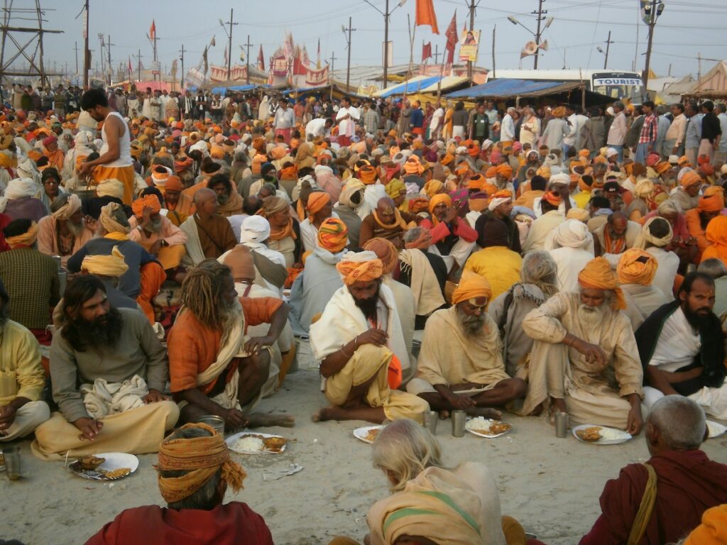 Large group of Hindu pilgrims sitting on the ground during Kumbh Mela, wearing traditional saffron robes and eating together.